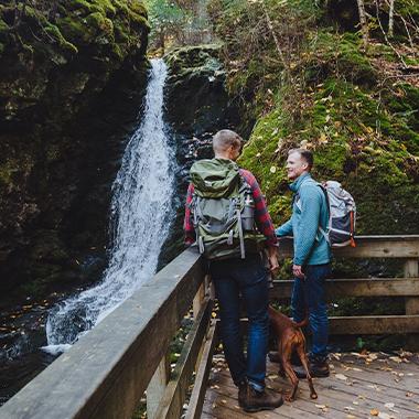 Two people standing on a deck in the forest, with a waterfall in the background.