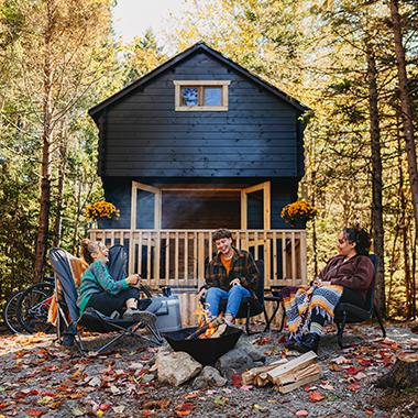 Three people sitting around a campfire in a forest with a cabin in the background.